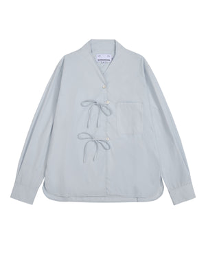 The DART COLLAR SHIRT by Sundae School is a light blue long-sleeve shirt crafted from a soft cotton-nylon blend, featuring two front tie closures, a single chest pocket, and a collarless neckline. Inspired by the relaxed silhouette of a shawl collar shirt, this men’s button-down is displayed flat against a white background.