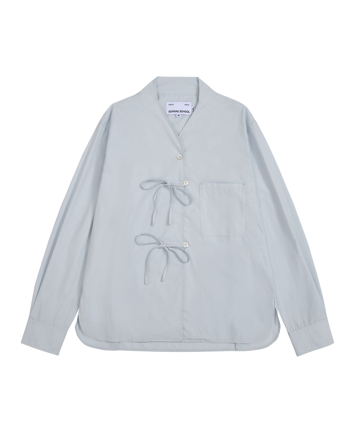 The DART COLLAR SHIRT by Sundae School is a light blue long-sleeve shirt crafted from a soft cotton-nylon blend, featuring two front tie closures, a single chest pocket, and a collarless neckline. Inspired by the relaxed silhouette of a shawl collar shirt, this men’s button-down is displayed flat against a white background.