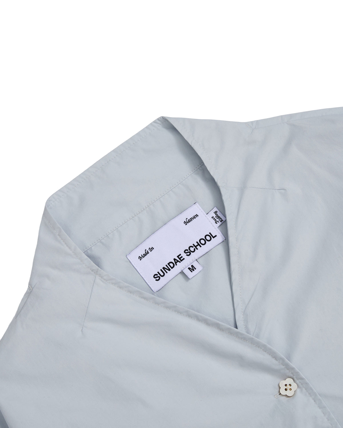 Here’s your rewritten sentence using the provided product data: The DART COLLAR SHIRT by Sundae School is a light blue button-down for men, featuring a white interior label that reads “Sundae School” and “Made in Korea.” Crafted from a comfortable cotton-nylon blend, this shirt is finished at the top with a small cream-colored button.