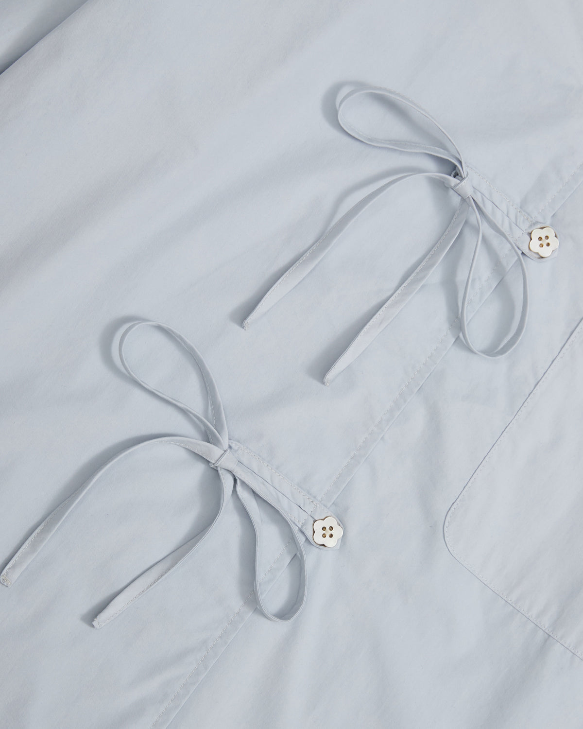 Close-up of Sundae School’s DART COLLAR SHIRT in light blue cotton-nylon blend, featuring two sewn bows and two four-hole white buttons arranged in a clean, minimal style reminiscent of a shawl collar shirt.
