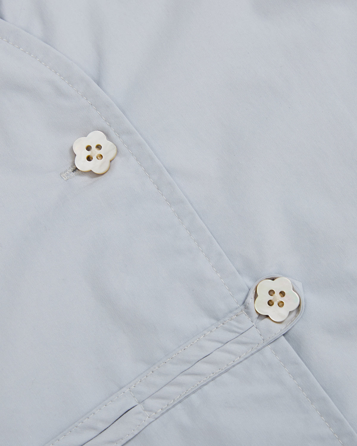 Close-up of a light blue cotton-nylon blend fabric from the Sundae School DART COLLAR SHIRT, featuring two white, four-hole flower-shaped buttons. The buttons are attached at intersecting stitched seams, adding subtle detail to this classic shirt design.