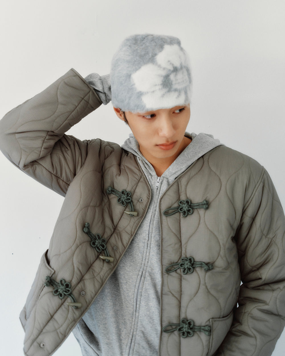 A person wearing a GREY DDUL BEANIE and a white floral pattern quilted jacket with traditional knotted closures over a grey hoodie.