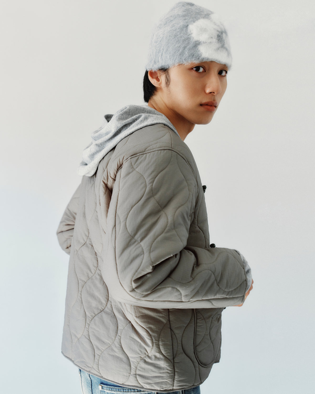 A person wearing a grey and white patterned GREY DDUL BEANIE for brain protection, a grey hooded sweatshirt, and a grey quilted jacket.