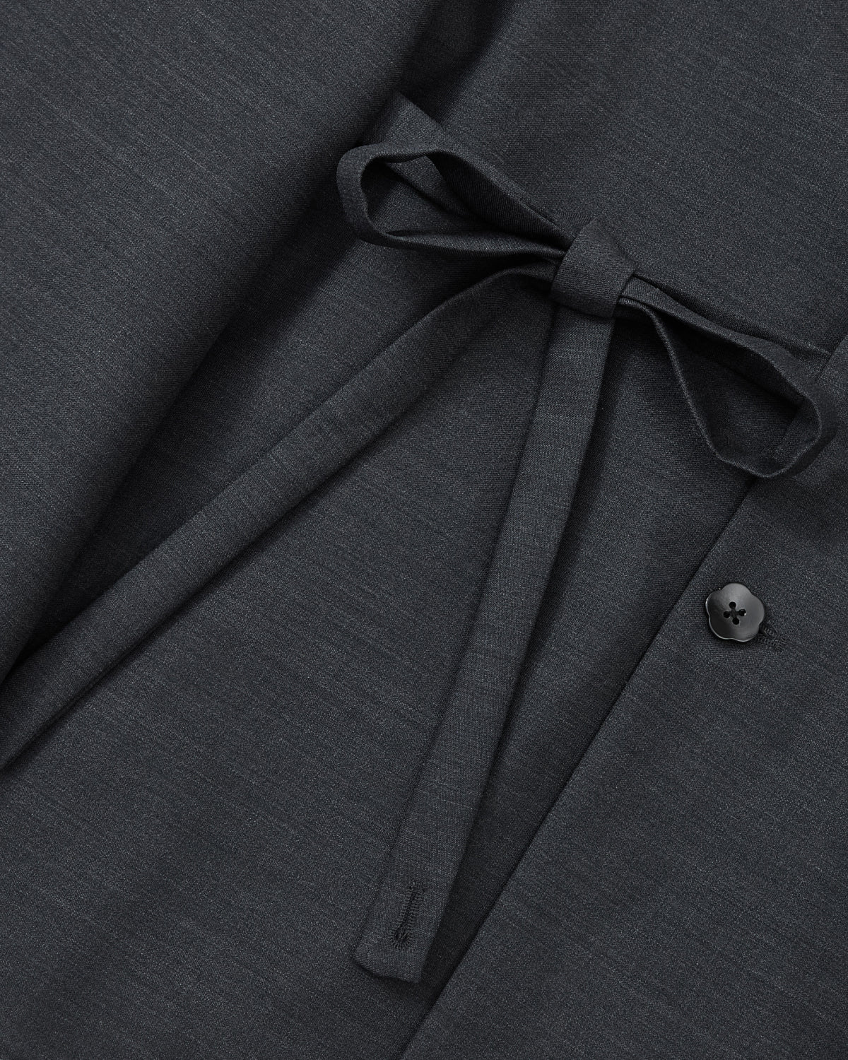 A close-up of the Sundae School CHARCOAL DOUBLE DUTY BLAZER highlights its dark gray premium wool blend, featuring a fabric belt tied in a bow for a subtle hanbok-inspired silhouette, with a single black button accenting the right side.