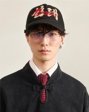 A person wears the Sundae School BLACK DRAGON CAP featuring a red and beige Korean dragon motif, clear eyeglasses, a black mandarin-collar jacket, white shirt, and red patterned tie against a plain background.