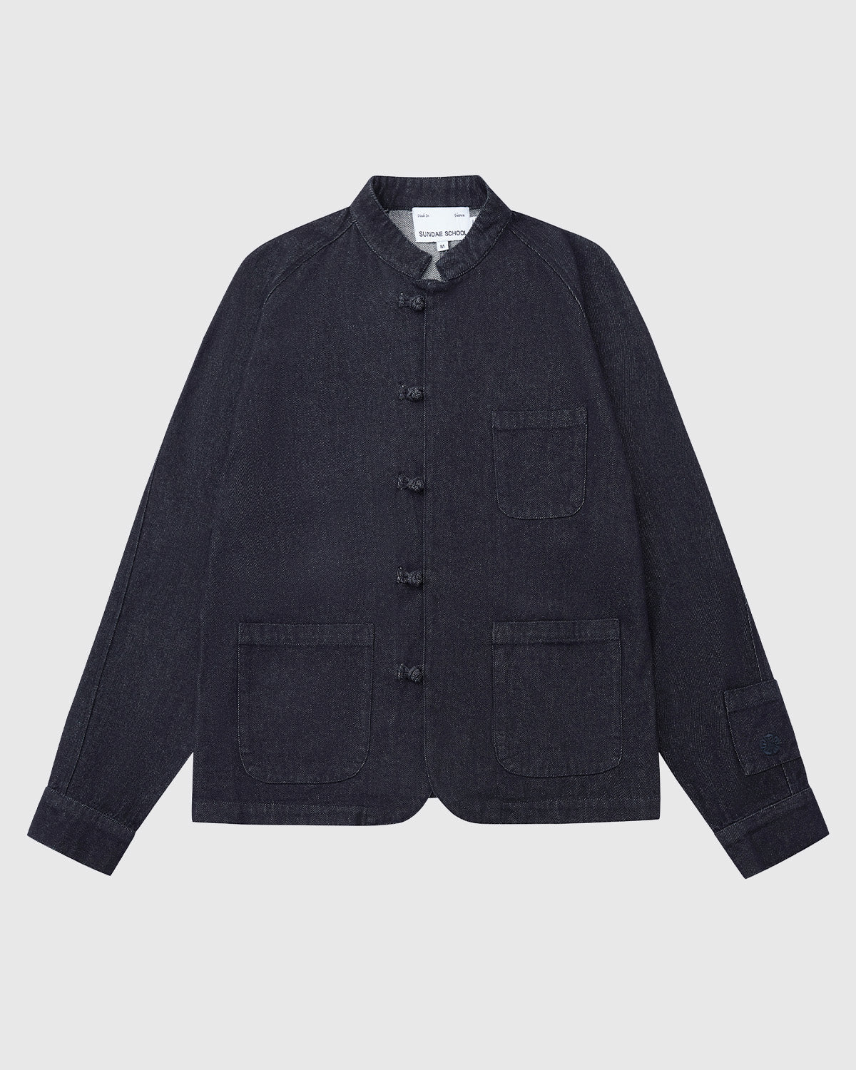 The Sundae School DENIM CHORE JACKET features dark denim, a mandarin collar, three front patch pockets, a button-up front, and long sleeves, shown laid flat on a white background.