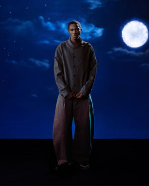 Against a nighttime backdrop with a bright full moon, a person wears the Sundae School BLACK CHINA COLLAR KNOT SHACKET and loose pants. The dim lighting adds to the moody atmosphere.