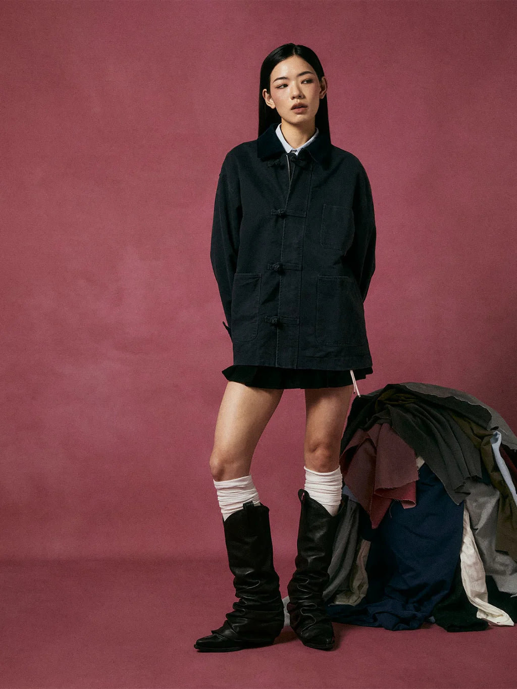 A model stands against a dusty rose studio backdrop wearing SUNDAE SCHOOL KNOT BARN JACKET over a white collared shirt, a black mini skirt, white knee-high socks, and black leather boots. A pile of folded garments sits to the right.