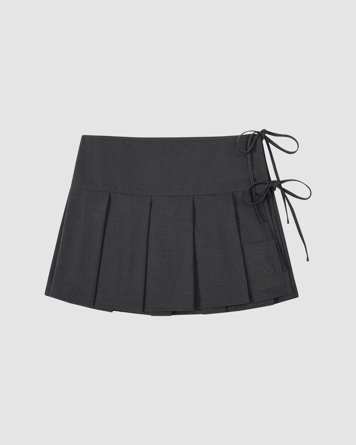 The FLARE SKIRT PANTS by Sundae School is a dark gray pleated mini skirt with two bows on the right and a rectangular patch near the hem, styled like apron skirt pants. The product is shown laid flat on a light surface.