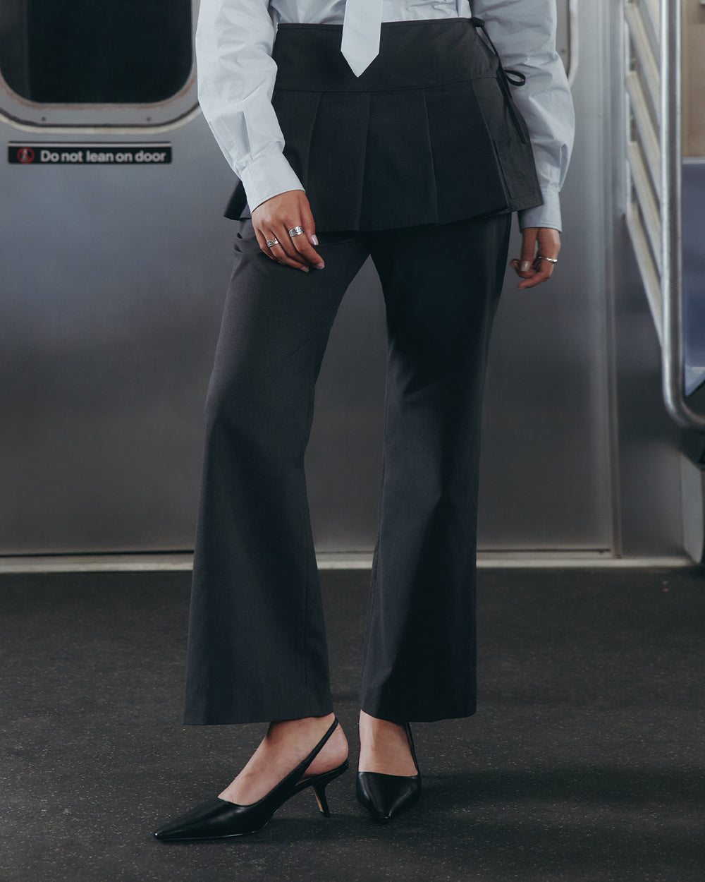 Dressed in Sundae School's FLARE SKIRT PANTS, a white shirt, and black heels, a person stands near the door inside a subway train. The summer wool trousers feature a removable pleated skirt. Their face is not visible.