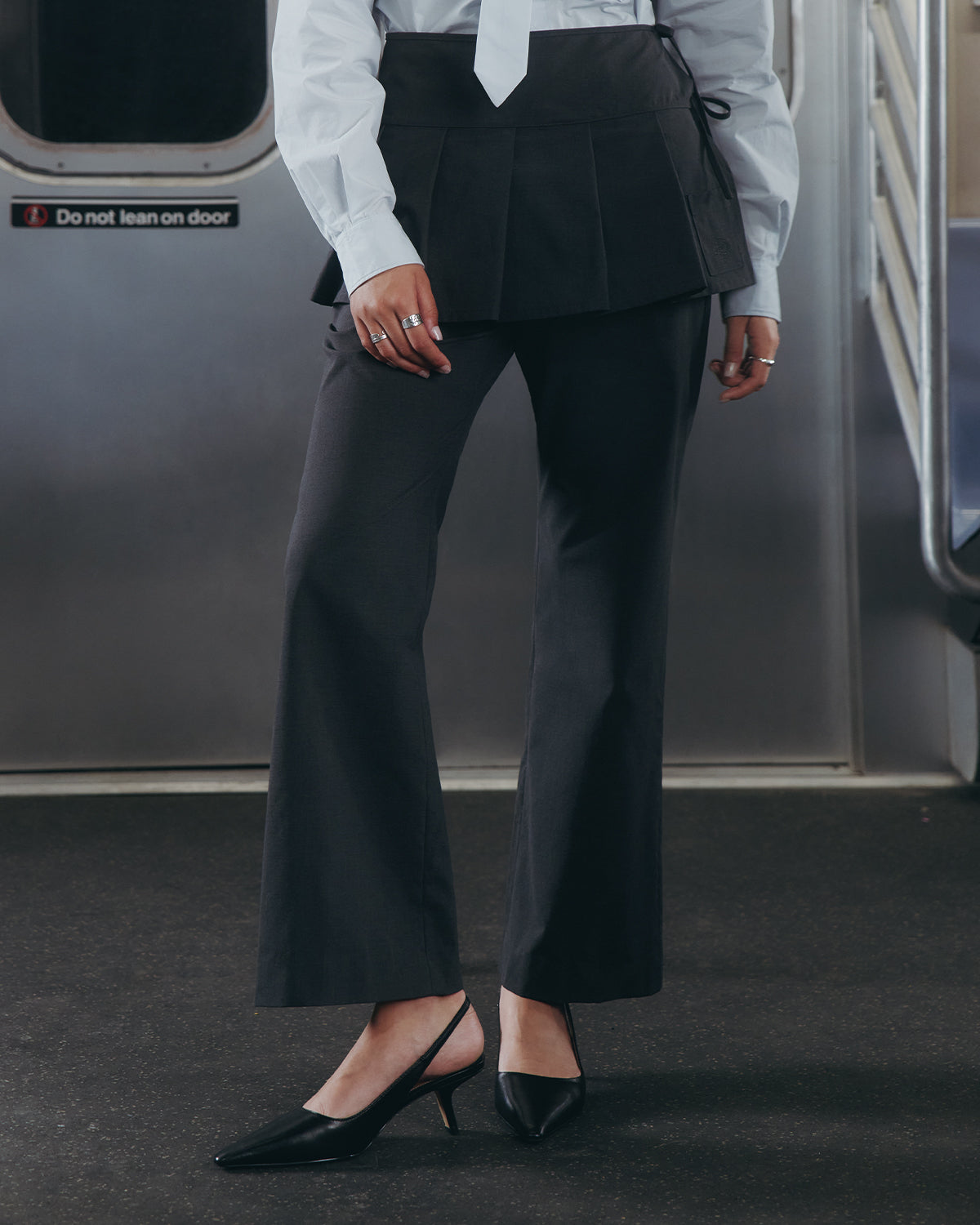 Dressed in Sundae School's FLARE SKIRT PANTS, a white shirt, and black heels, a person stands near the door inside a subway train. The summer wool trousers feature a removable pleated skirt. Their face is not visible.