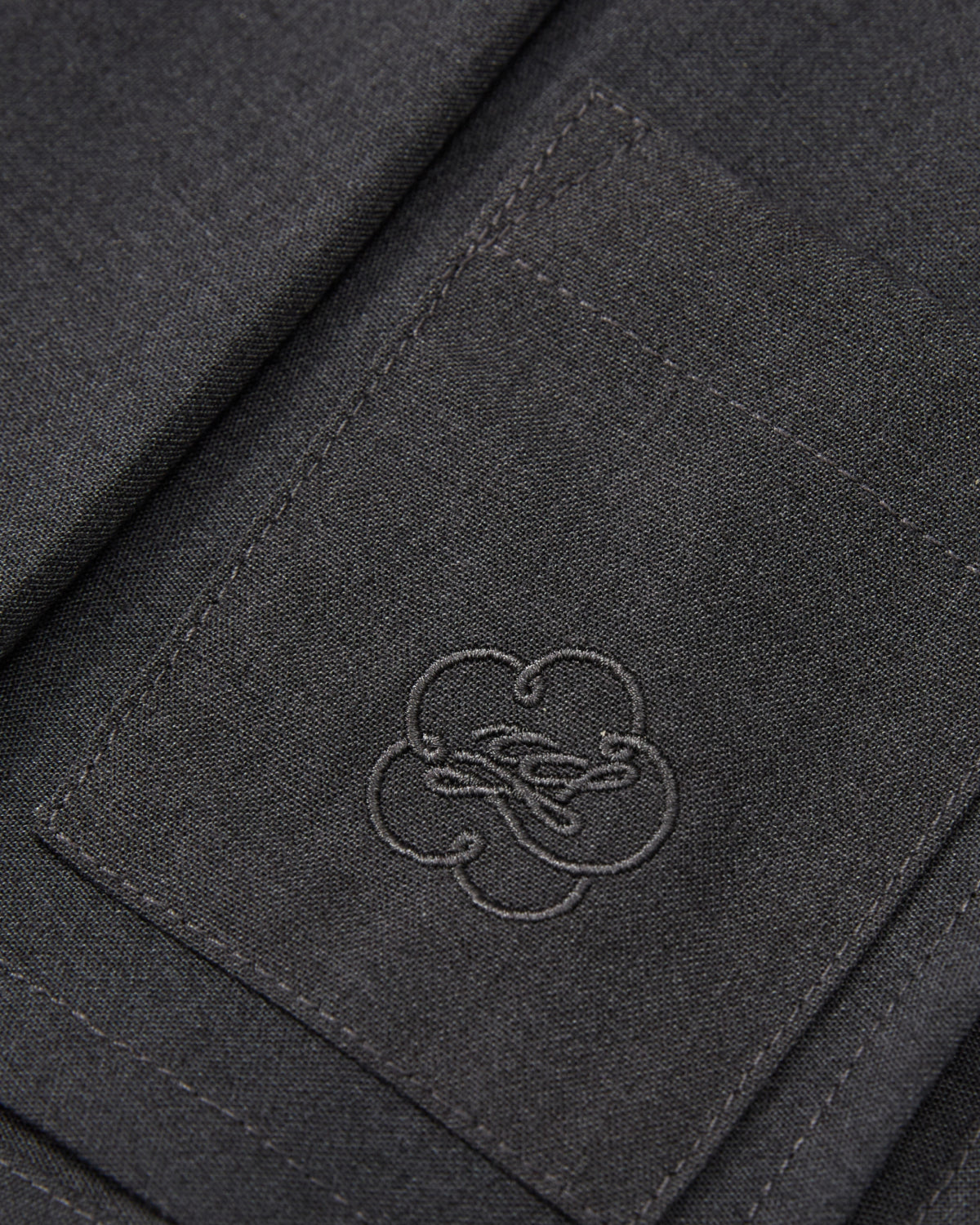 Close-up of a dark gray fabric pocket with detailed stitching and an embroidered clover-shaped logo in the center, featured on the FLARE SKIRT PANTS by Sundae School.