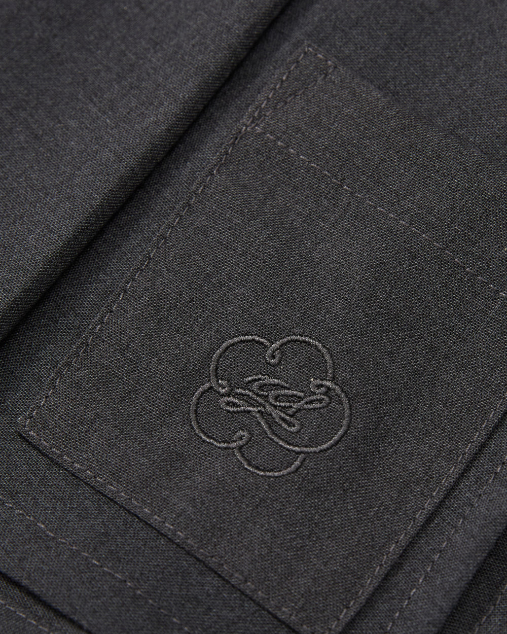 Close-up of a dark gray fabric pocket with detailed stitching and an embroidered clover-shaped logo in the center, featured on the FLARE SKIRT PANTS by Sundae School.