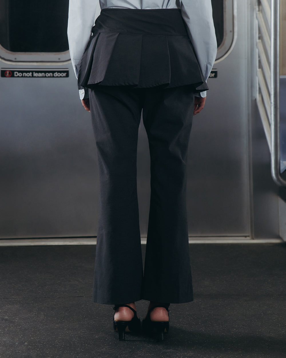 A person stands on a subway train wearing Sundae School FLARE SKIRT PANTS, paired with a light blue shirt and black heels. Subway doors and a metal railing are visible in the background.