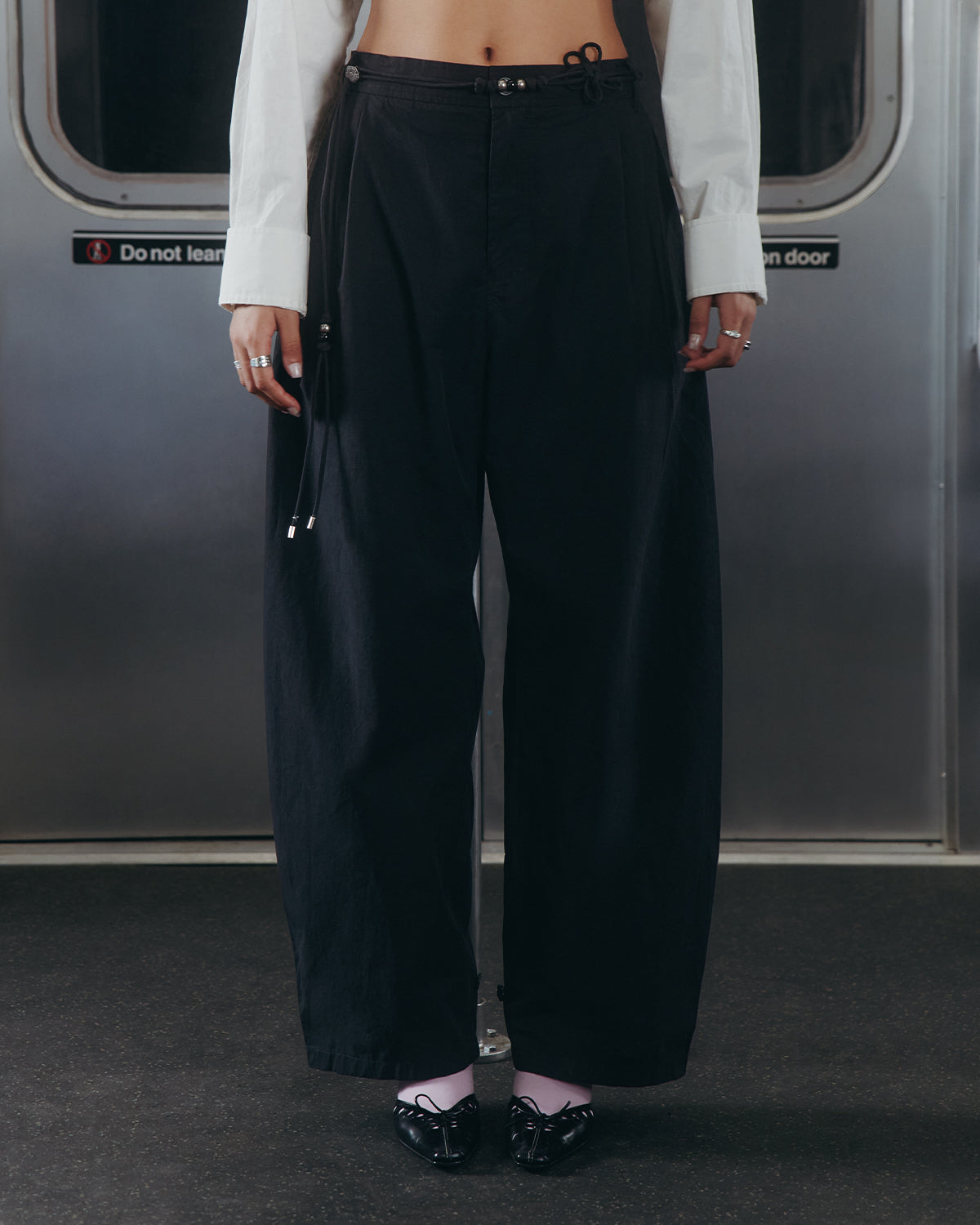 A person stands in a subway car wearing Sundae School’s BLACK FLOWER NORIGAE LINEN TROUSERS, paired with a white shirt, white socks, and black ballet flats. Their hands rest at their sides, with the subway door and warning sign visible in the background.