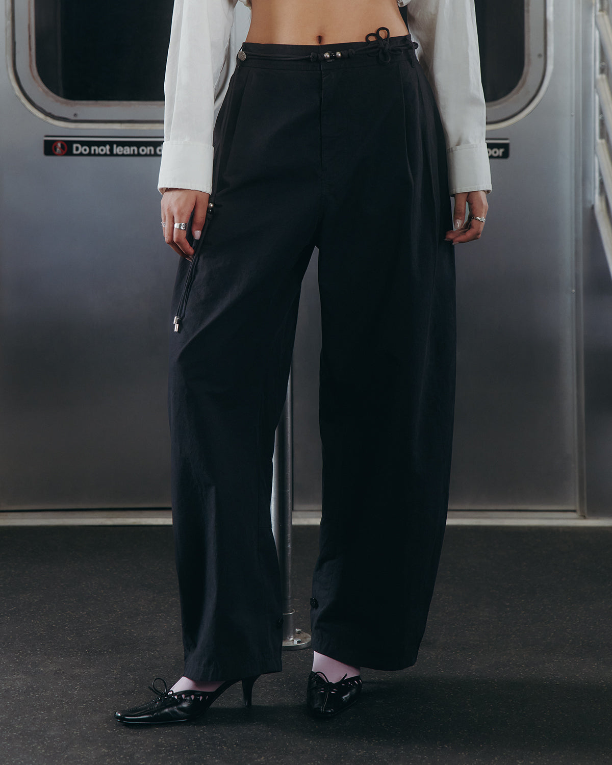 A person stands in a subway car wearing Sundae School's BLACK FLOWER NORIGAE LINEN TROUSERS paired with a white shirt, black heels, and pale socks. Their head is out of frame, and a sign behind them reads, 