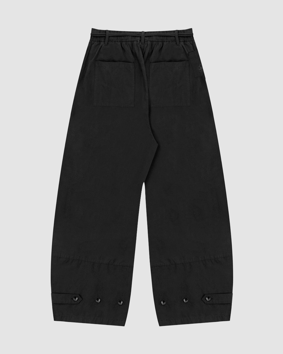The BLACK FLOWER NORIGAE LINEN TROUSERS by Sundae School, crafted from black cotton-linen with two back pockets, a discreet stash pocket, adjustable buttoned ankle straps, and an elastic waistband, are displayed against a plain light gray background.