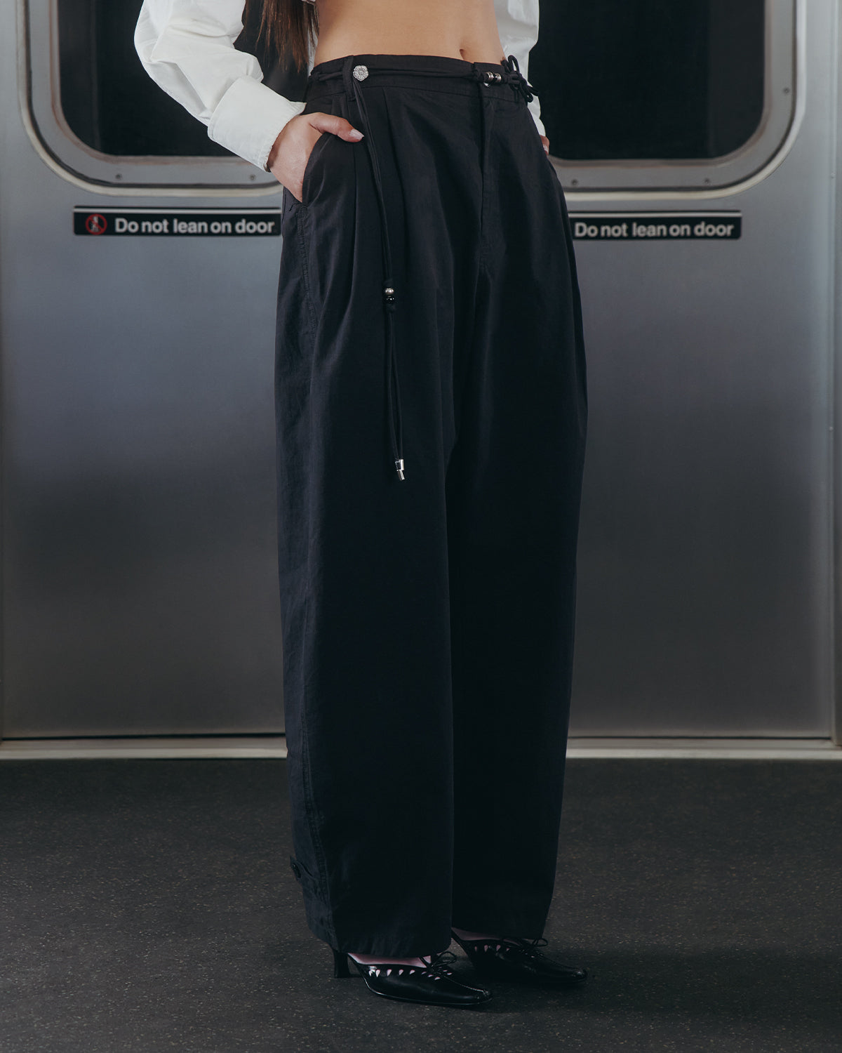A person wearing Sundae School's BLACK FLOWER NORIGAE LINEN TROUSERS and black shoes stands in front of a subway door with "Do not lean on door" signs visible; their upper body is out of frame.