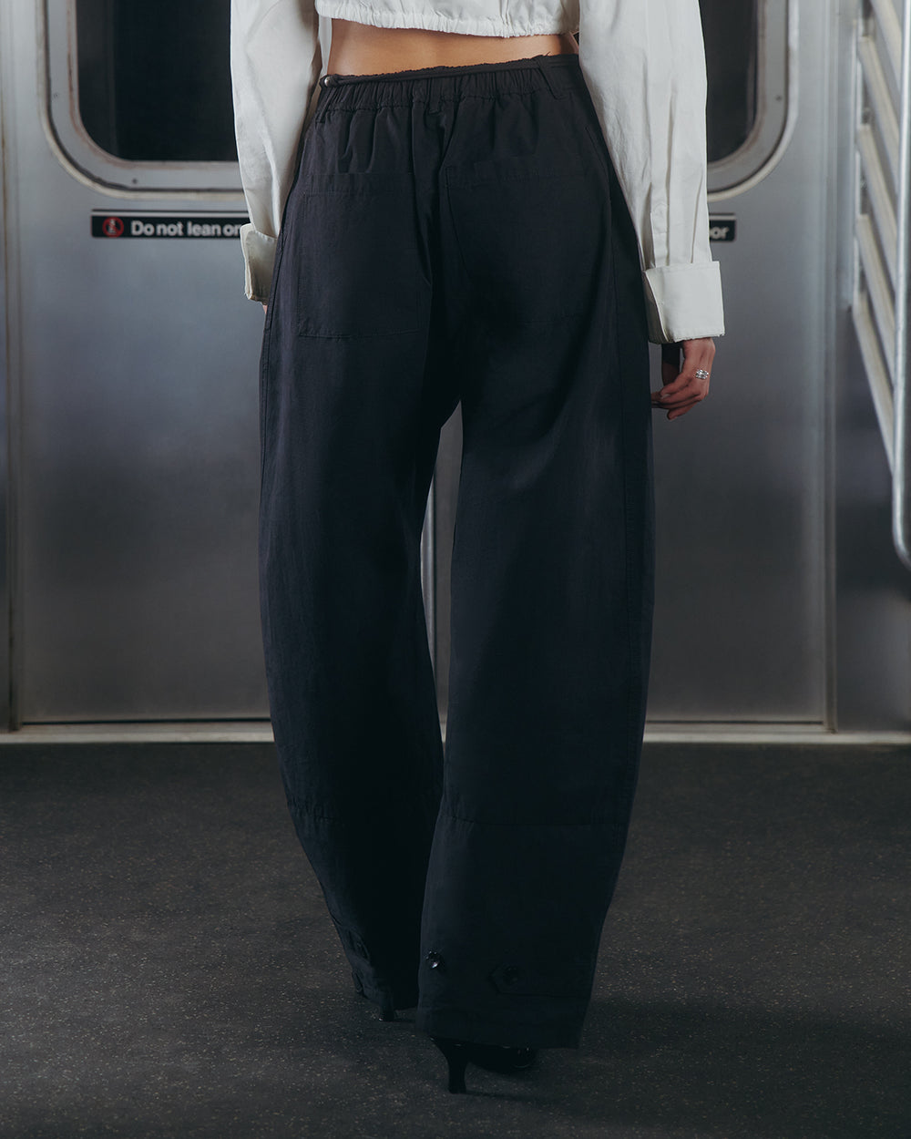 A person wearing Sundae School’s BLACK FLOWER NORIGAE LINEN TROUSERS and a white shirt stands with their back to the camera inside a subway train, near closed silver doors and a metal handrail.