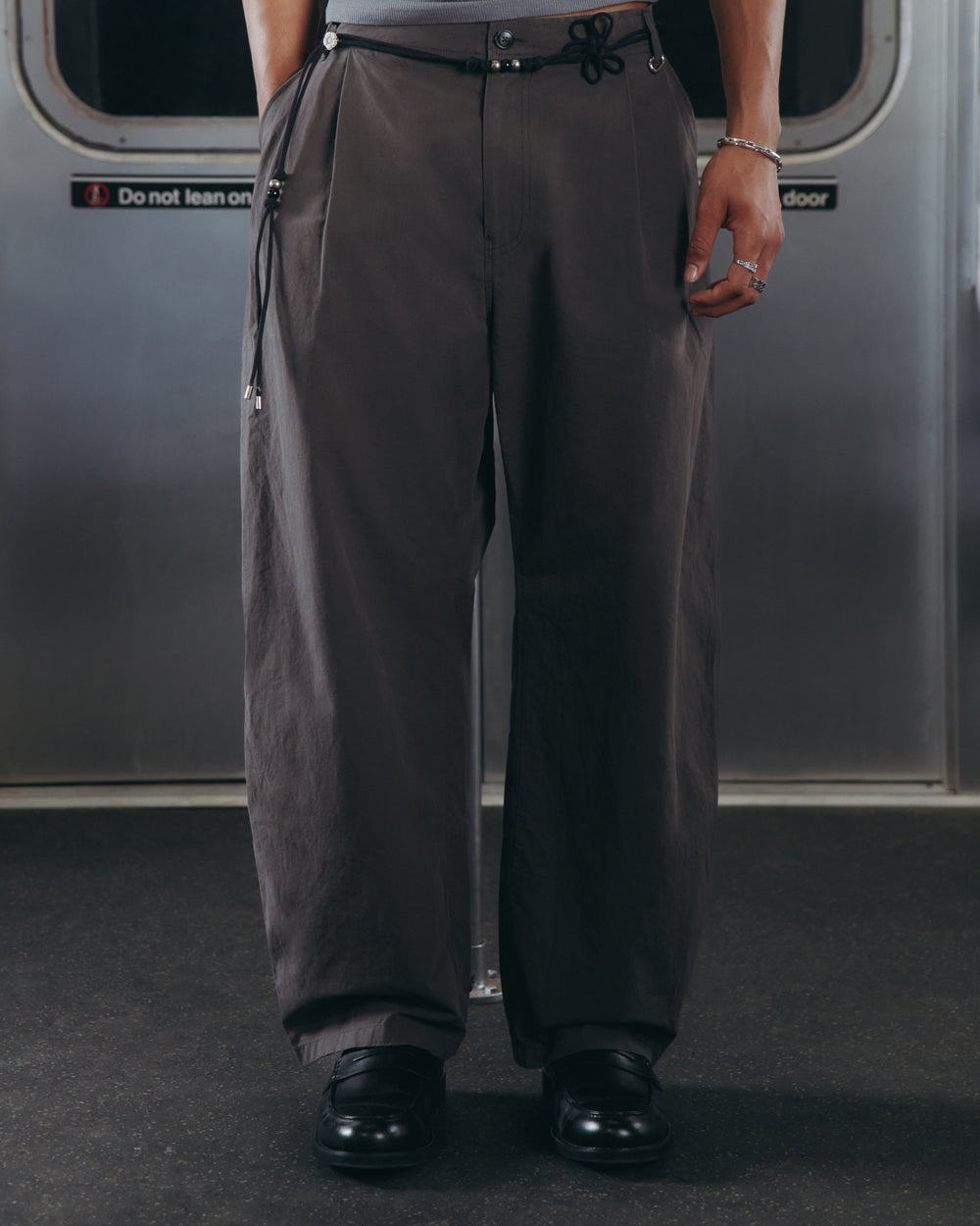 A person wearing Sundae School’s CHARCOAL FLOWER NORIGAE LINEN TROUSERS—loose, dark gray pants with a black drawstring—paired with black shoes is standing on a subway train, with only the lower half of their body visible.
