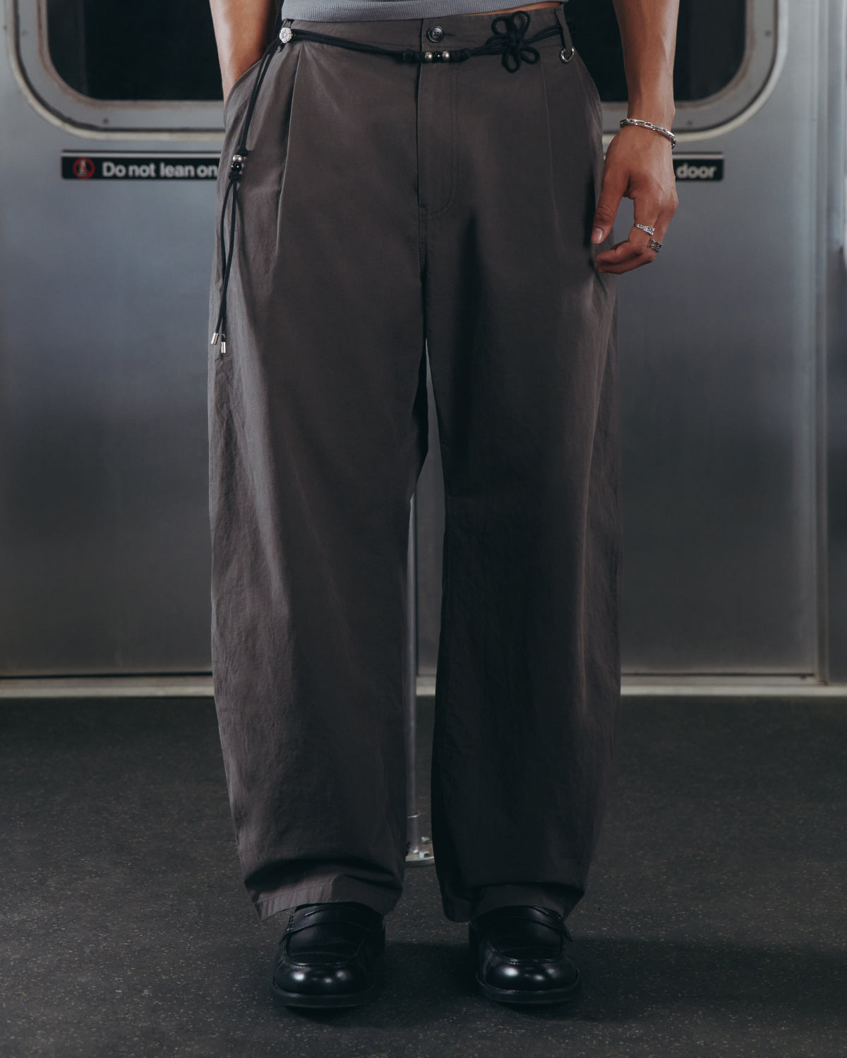 A person wearing Sundae School’s CHARCOAL FLOWER NORIGAE LINEN TROUSERS—loose, dark gray pants with a black drawstring—paired with black shoes is standing on a subway train, with only the lower half of their body visible.