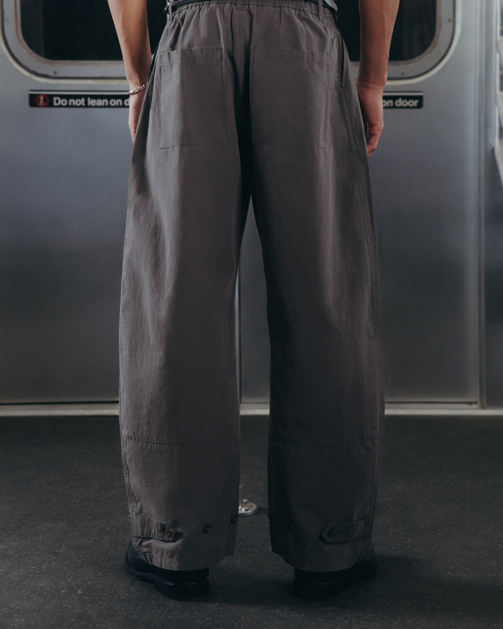 Wearing Sundae School's CHARCOAL FLOWER NORIGAE LINEN TROUSERS, a person stands in wide-legged gray pants on public transit, facing away from the camera. A sign on the door behind reads Do not lean on door.