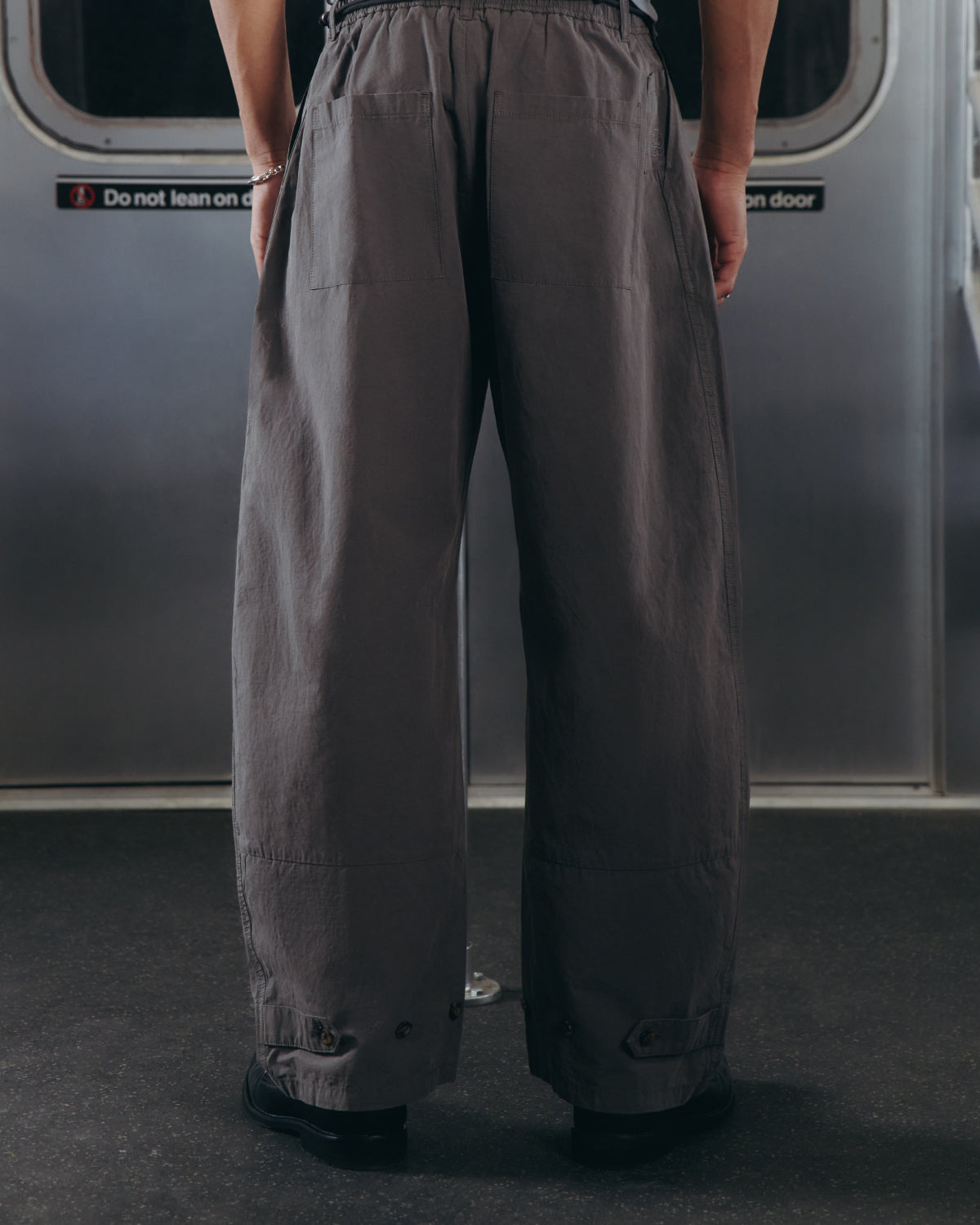 Wearing Sundae School's CHARCOAL FLOWER NORIGAE LINEN TROUSERS, a person stands in wide-legged gray pants on public transit, facing away from the camera. A sign on the door behind reads Do not lean on door.