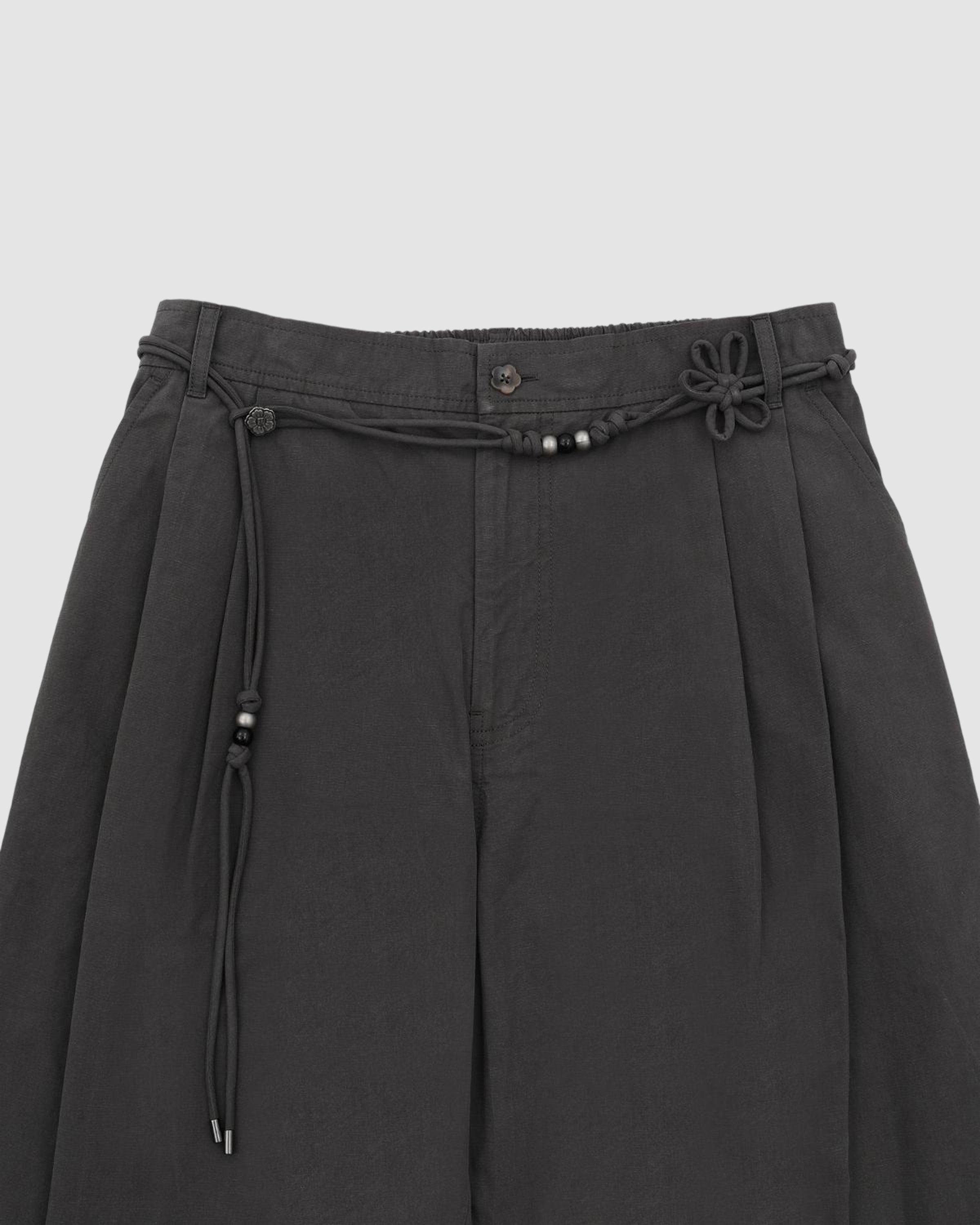 Introducing the CHARCOAL FLOWER NORIGAE LINEN TROUSERS by Sundae School: Korean-inspired black pleated pants made from soft cotton linen balloon fabric, featuring a detachable norigae rope belt adorned with a decorative knot and small black beads—beautifully displayed against a plain light background.