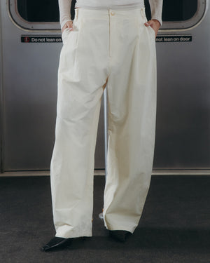 A person stands in front of a subway door with “Do not lean on door” signs, hands in pockets and upper body partially out of frame, wearing Sundae School’s IVORY FLOWER NORIGAE LINEN TROUSERS—loose, high-waisted cream-colored pants—paired with black pointed shoes.