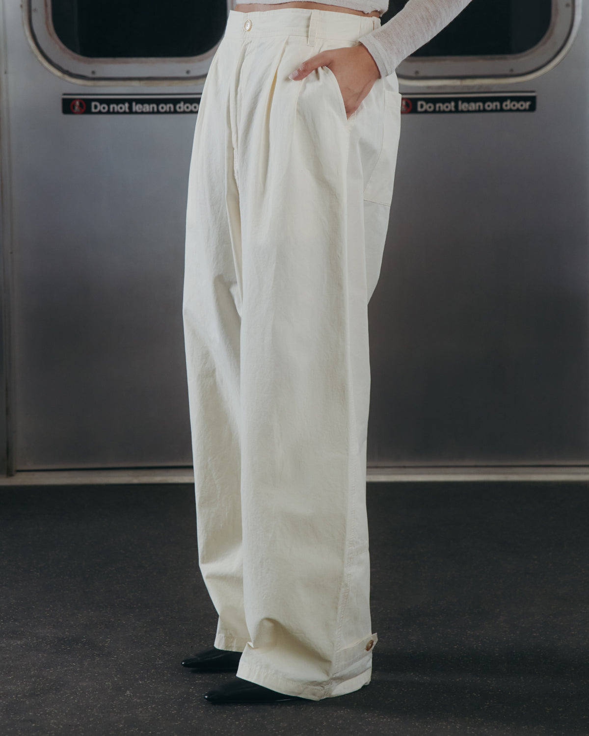 A person wearing Sundae School’s IVORY FLOWER NORIGAE LINEN TROUSERS stands in a subway car with hands in pockets, framed by metal subway doors featuring 