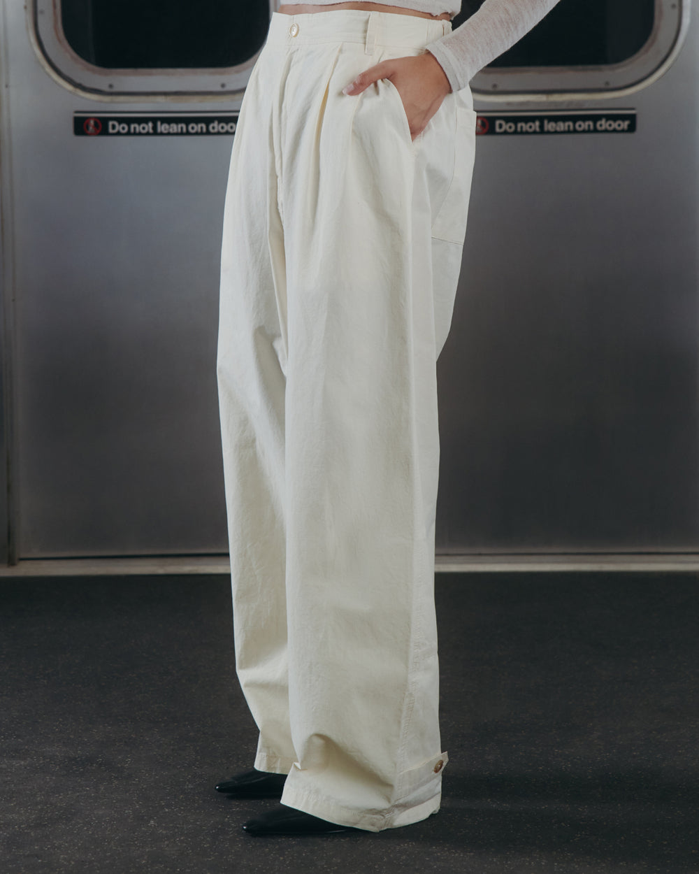 A person wearing Sundae School’s IVORY FLOWER NORIGAE LINEN TROUSERS stands in a subway car with hands in pockets, framed by metal subway doors featuring "Do not lean on door" signs.