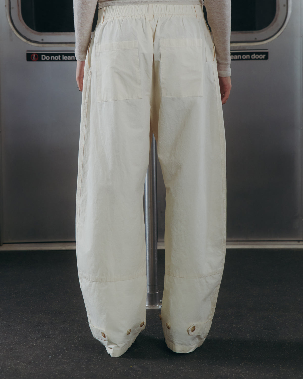A person wearing Sundae School's IVORY FLOWER NORIGAE LINEN TROUSERS stands on public transportation, facing away. The background features a metal door with a sign that reads, "Do not lean on door.