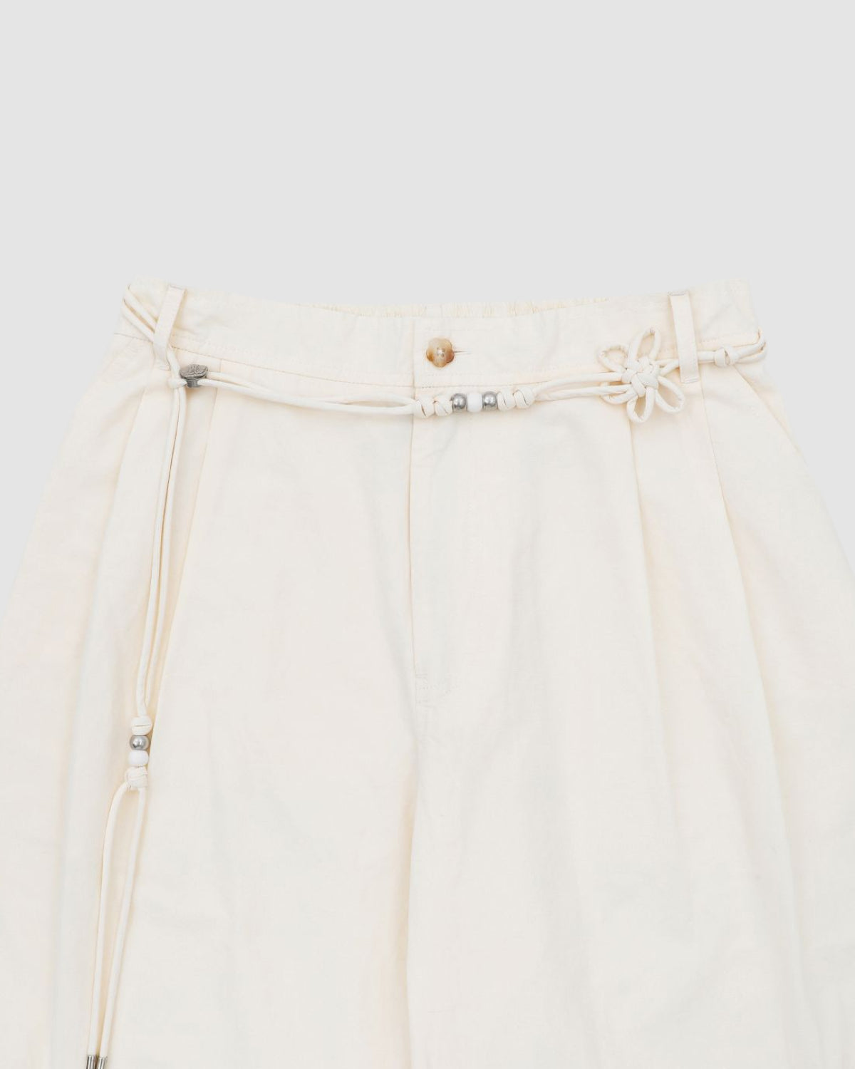 The IVORY FLOWER NORIGAE LINEN TROUSERS by Sundae School are cream-colored, high-waisted pants crafted from a cotton-linen blend. They feature a button closure and a detachable norigae rope belt adorned with silver beads and a flower-shaped knot, drawing inspiration from traditional Korean heritage pants. These stylish balloon trousers are set against a light gray background.