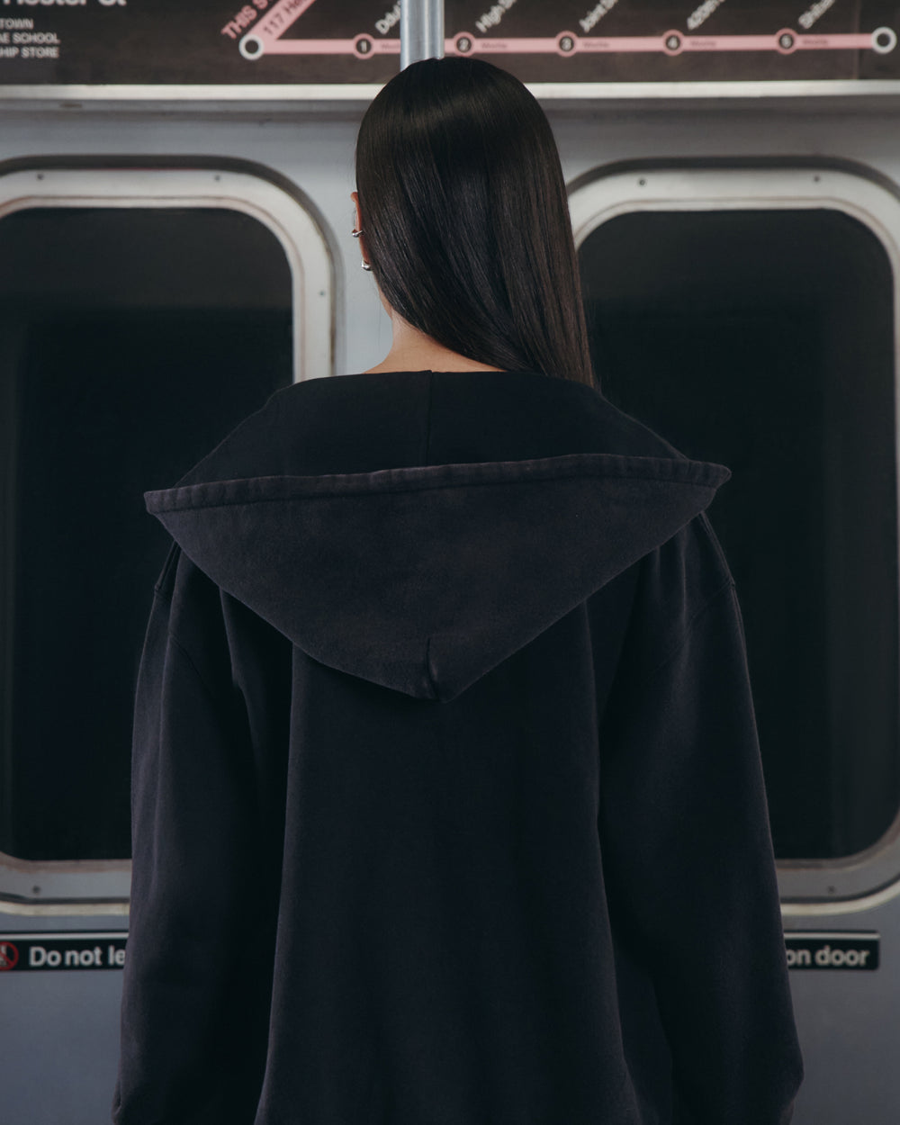 A person with long, straight hair stands inside a subway train, facing away from the camera and gazing out the window. They are wearing Sundae School's BLACK WASH FLOWER KNOT ZIP UP hoodie, distinguished by a Maehwa knot zipper pull and subtle Hangul Sundae School embroidery on the back. A subway map and safety sign can be seen in the background.