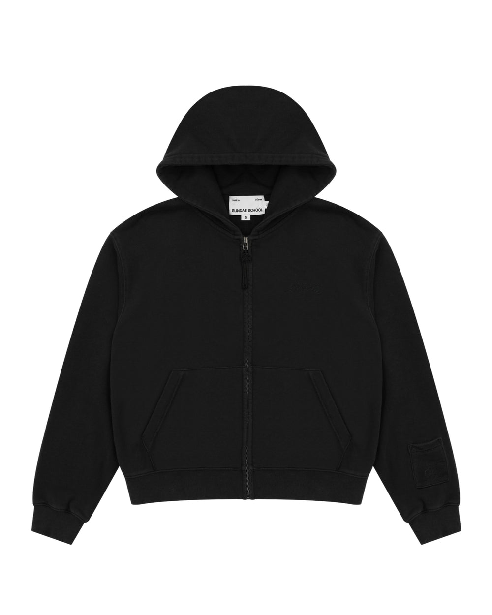 The BLACK WASH FLOWER KNOT ZIP UP by Sundae School is a plain black cotton zip-up hoodie with a hood, front pockets, and a small patch pocket on the left sleeve. It features a distinctive Maehwa knot zipper pull and is displayed laid flat on a white background.