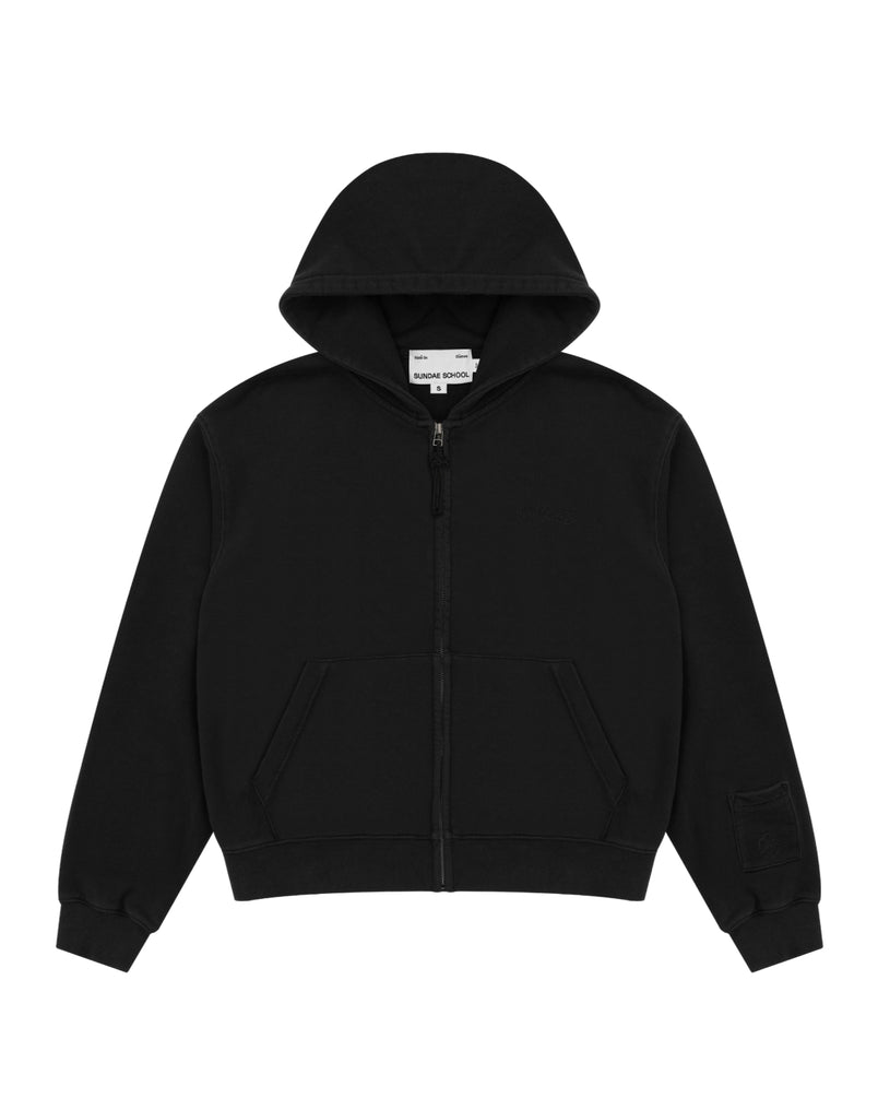 The BLACK WASH FLOWER KNOT ZIP UP by Sundae School is a plain black cotton zip-up hoodie with a hood, front pockets, and a small patch pocket on the left sleeve. It features a distinctive Maehwa knot zipper pull and is displayed laid flat on a white background.