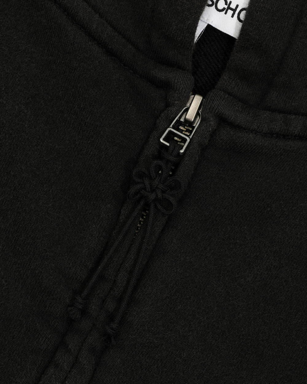 Close-up of the BLACK WASH FLOWER KNOT ZIP UP by Sundae School, highlighting the black cotton fabric, zipper with a Maehwa knot pull, and drawstrings. The top of the zipper is partially open, revealing a glimpse of a white tag with black text inside the collar.