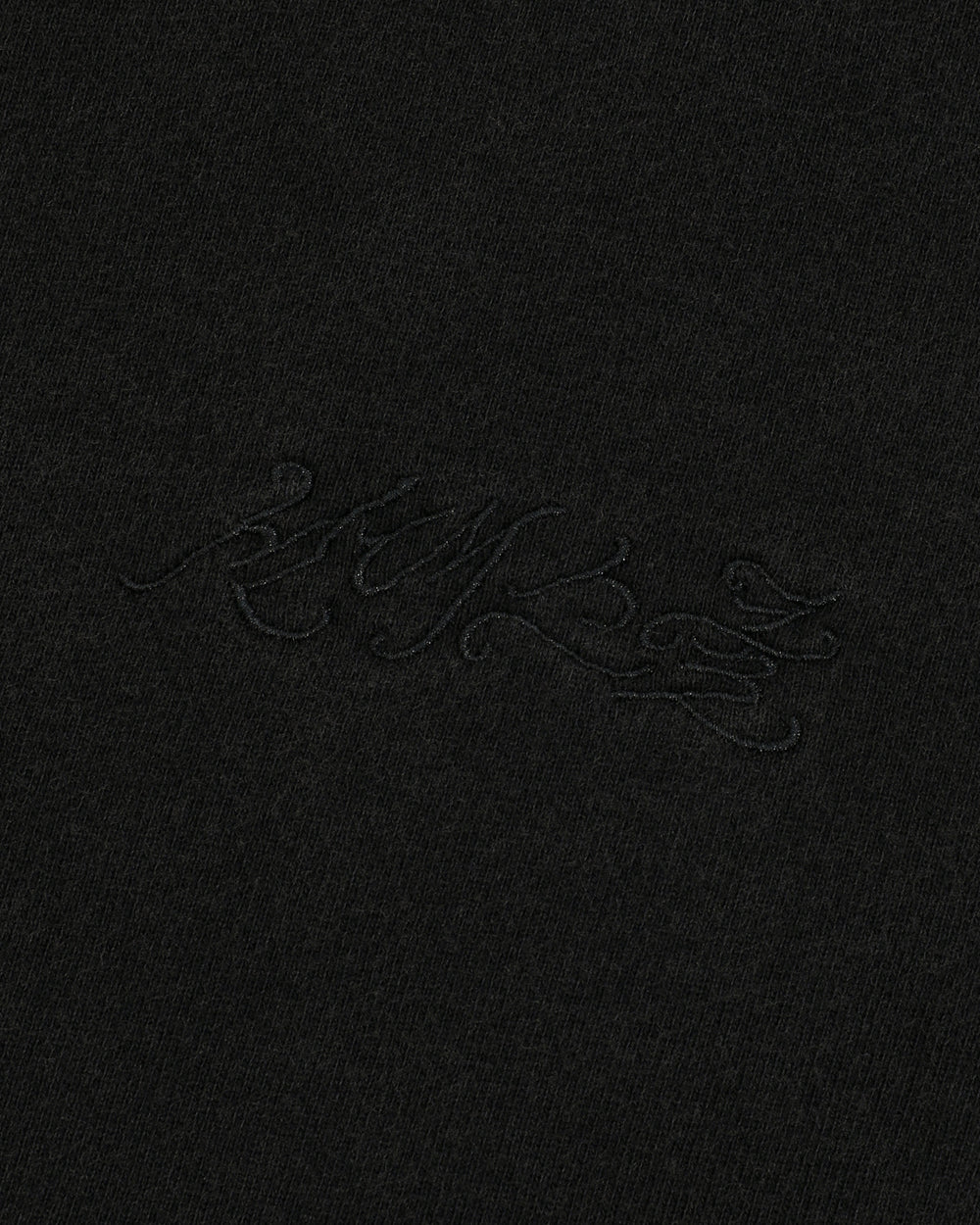 A close-up of black fabric featuring subtle, decorative black embroidered script that blends seamlessly with the material highlights the unique Hangul Sundae School embroidery on the BLACK WASH FLOWER KNOT ZIP UP hoodie by Sundae School.