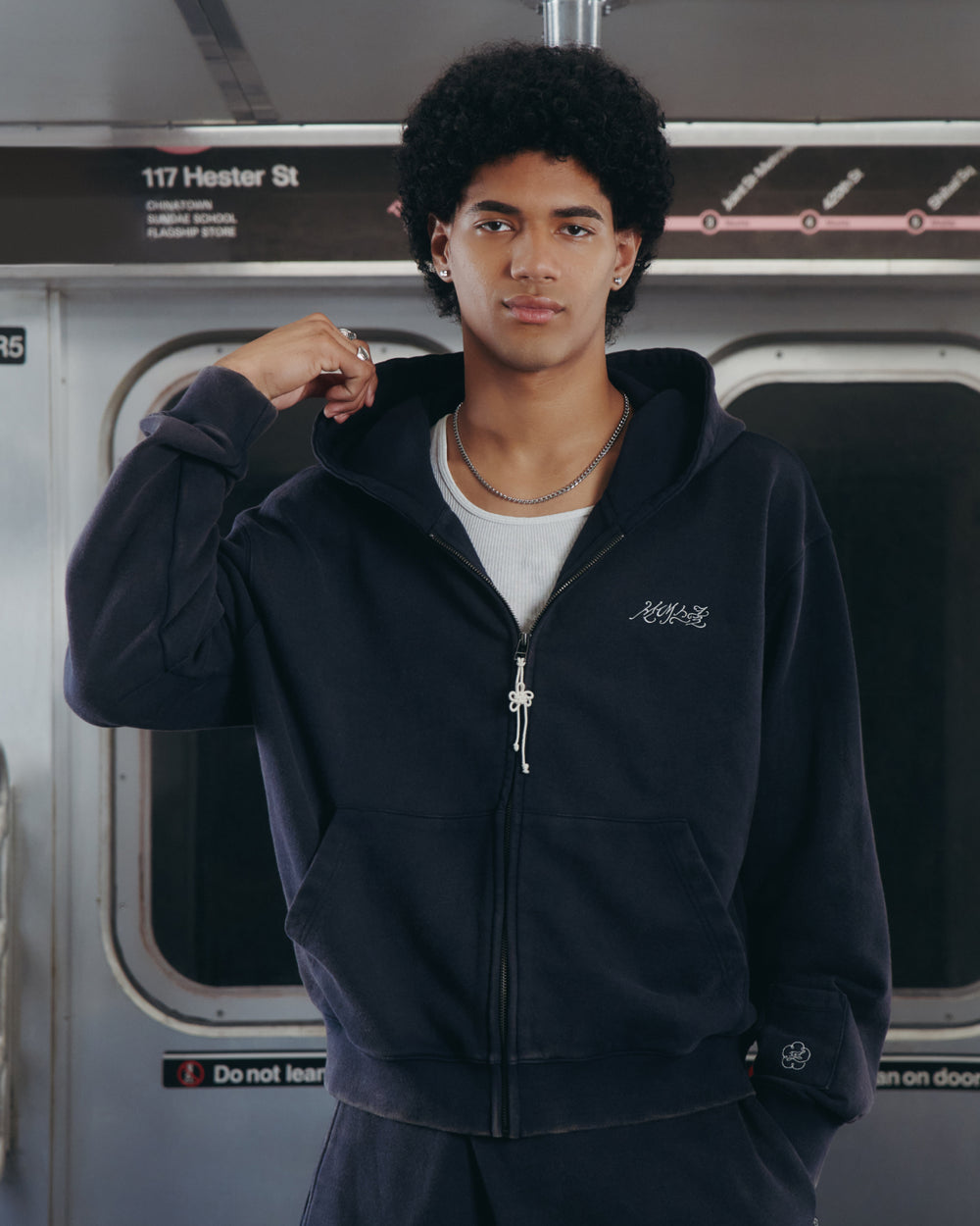 A young man with curly hair wearing the Sundae School NAVY WASH FLOWER KNOT ZIP UP hoodie and a white shirt stands in a subway train, holding his hood, with the "Hester St" station sign visible behind him. Subtle details, like the Maehwa knot accent on his hoodie, hint at his connection to Sundae School.