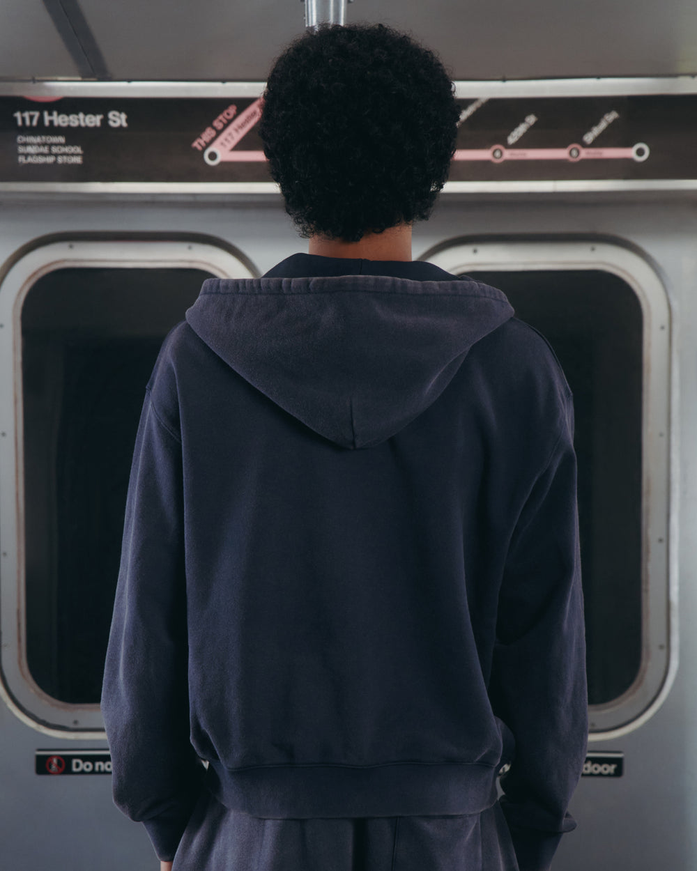 A person with short curly hair, wearing the Sundae School NAVY WASH FLOWER KNOT ZIP UP, stands facing away inside a subway train. A subway map and a "117 Hester St" sign are visible above the windows, hinting at destinations related to Hangul Sundae School.