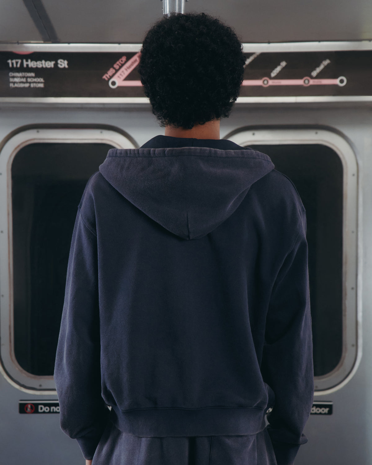 A person with short curly hair, wearing the Sundae School NAVY WASH FLOWER KNOT ZIP UP, stands facing away inside a subway train. A subway map and a "117 Hester St" sign are visible above the windows, hinting at destinations related to Hangul Sundae School.