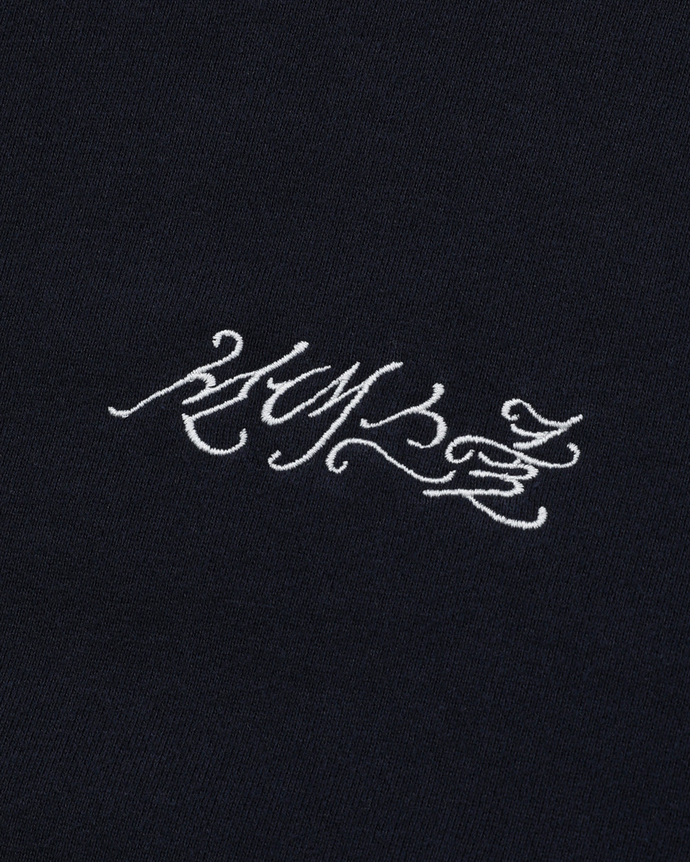 White calligraphic text is embroidered on a dark cotton background, reminiscent of the ornate lettering associated with Sundae School. The script is stylized and intricate, lending an enigmatic appeal to the NAVY WASH FLOWER KNOT ZIP UP hoodie by Sundae School.