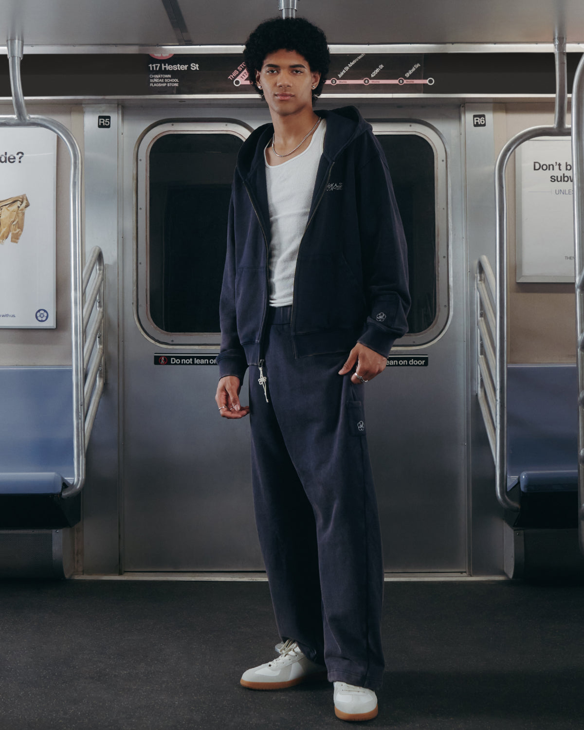 A person stands in a subway car wearing the Sundae School NAVY WASH FLOWER KNOT ZIP UP, matching pants, a white T-shirt, and white sneakers. The subway seats are empty, and the train map and informational signs—including references to Hangul Sundae School—are visible in the background.