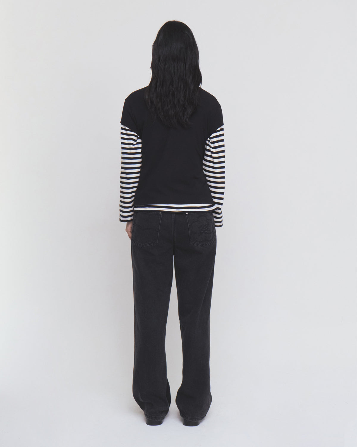 A person stands with their back to the camera against a plain white background, wearing Sundae School’s BLACK WIDOW FRESHMAN FIT DENIM and a black long-sleeve top featuring black and white striped sleeves.