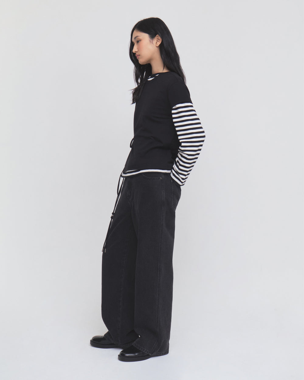 A person with long dark hair stands sideways against a plain white background, wearing Sundae School’s BLACK WIDOW FRESHMAN FIT DENIM jeans, paired with a black top layered over a black-and-white striped shirt and black shoes.