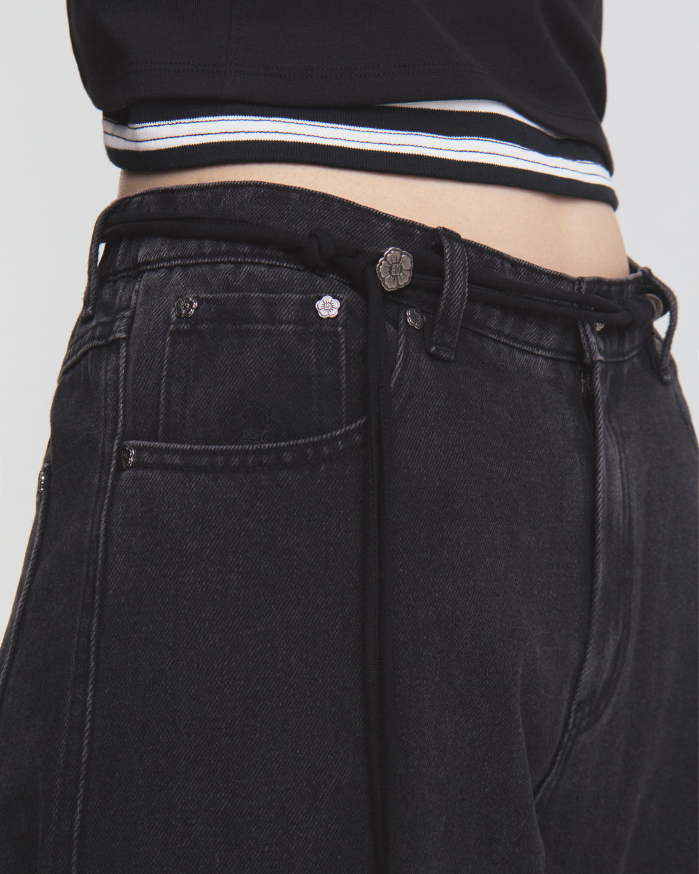 A close-up of a person wearing the Sundae School BLACK WIDOW FRESHMAN FIT DENIM, featuring high-waisted, black Hanbok-inspired jeans in a relaxed straight-leg fit with a Norigae belt. The look is paired with a black top accented by a white striped hem.