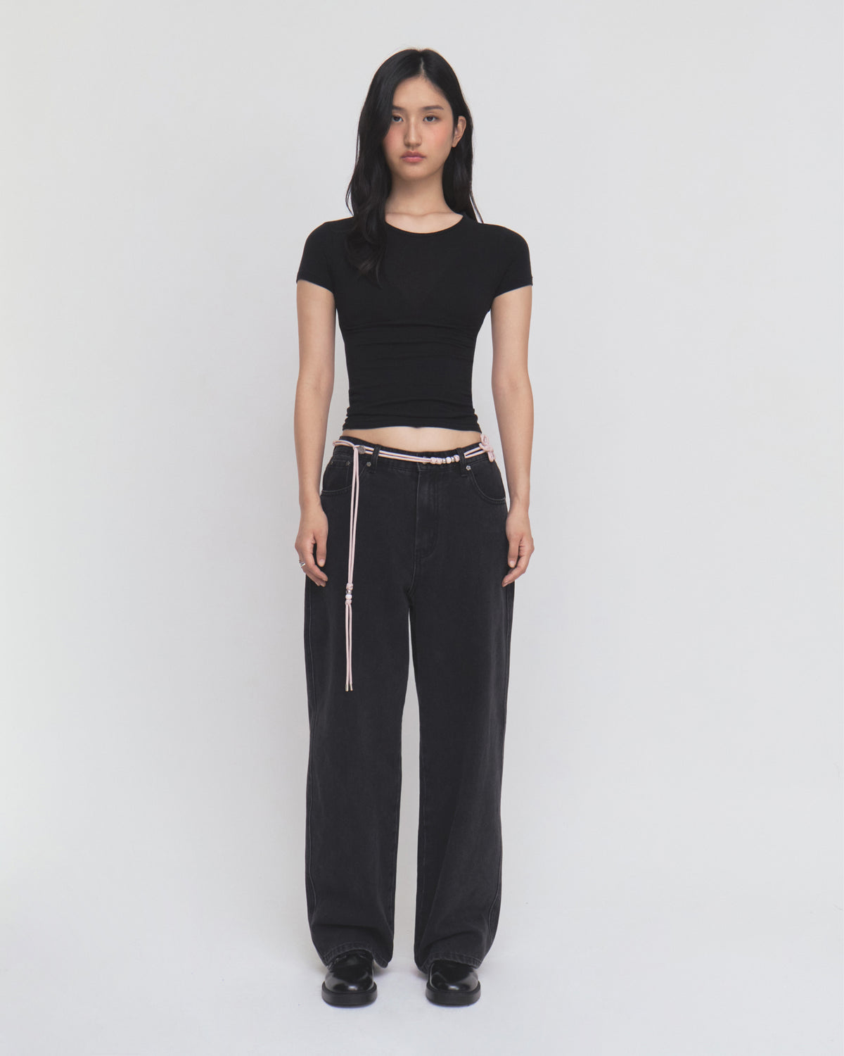 A woman stands against a plain white background, wearing a fitted black short-sleeve top and Sundae School BLACK WIDOW FRESHMAN FIT DENIM jeans styled with a delicate Norigae belt, paired with black shoes. She has long dark hair and a neutral expression.