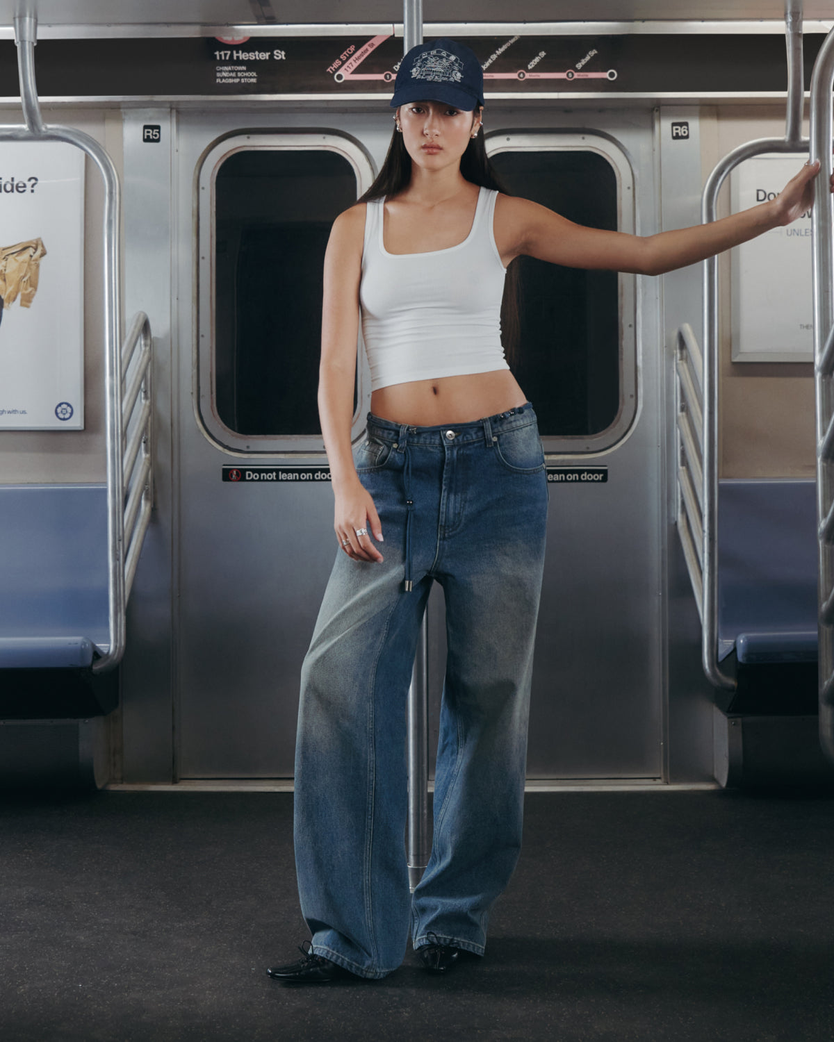 A woman in a white tank top, Sundae School MEDIUM BLUE FRESHMAN FIT DENIM, and a blue cap stands in a subway car, resting one arm on a pole with subway doors and empty seats behind her.