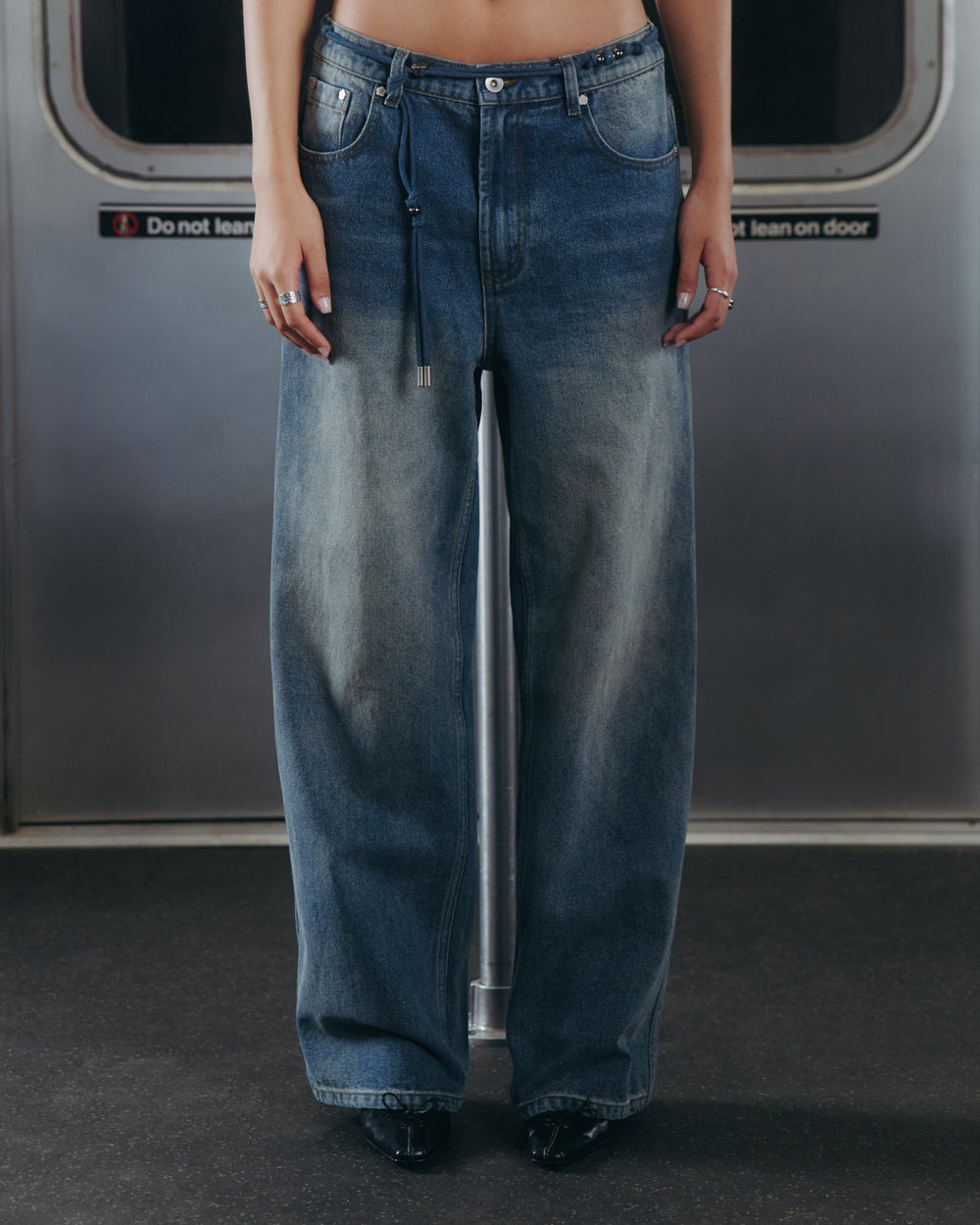 A person wearing Sundae School’s BLUE DREAM FRESHMAN FIT DENIM jeans and black shoes stands in front of a subway door with a sign that reads, “Do not lean on door.” Only the lower torso and legs are visible in the image.