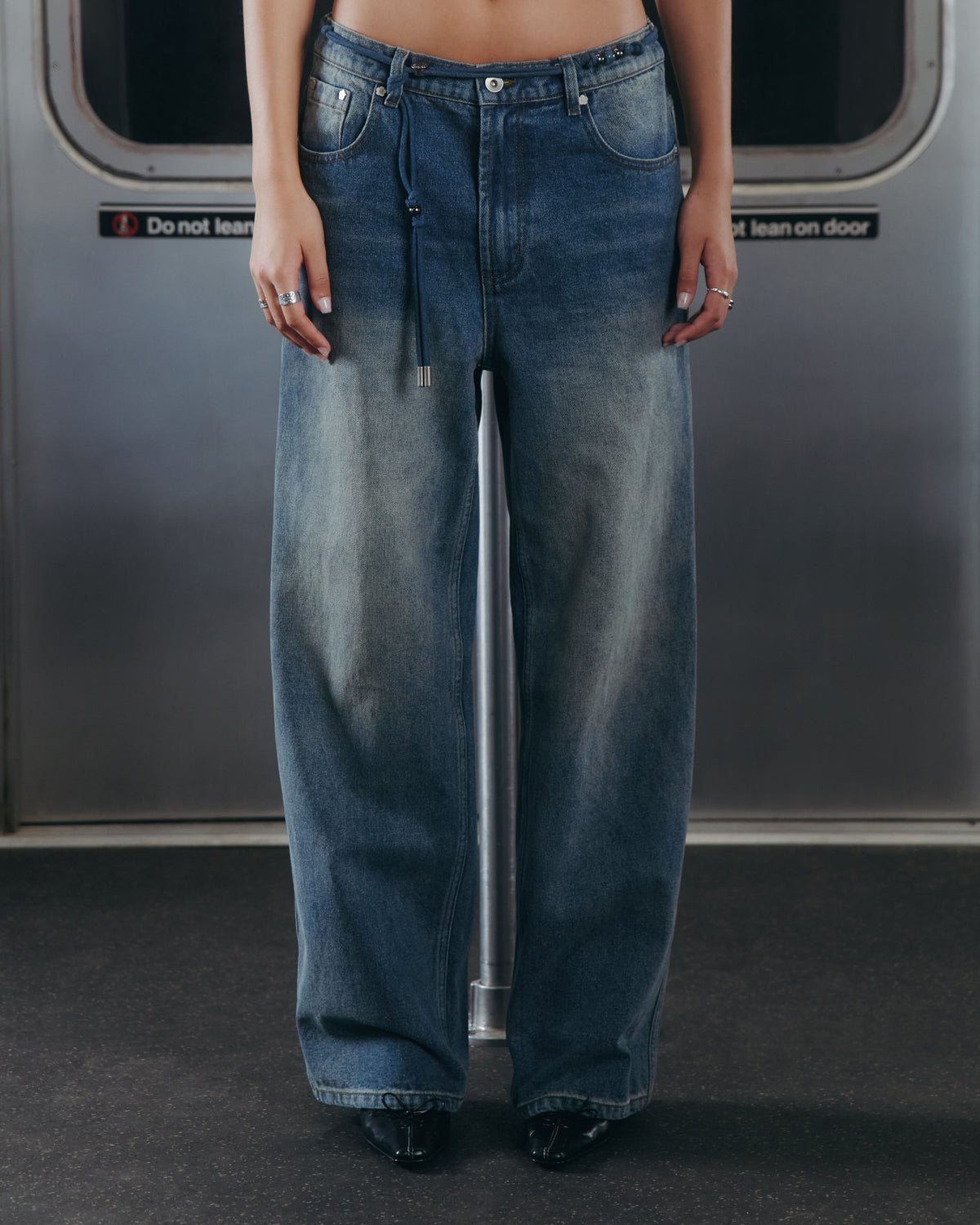 A person wearing Sundae School’s BLUE DREAM FRESHMAN FIT DENIM jeans and black shoes stands in front of a subway door with a sign that reads, “Do not lean on door.” Only the lower torso and legs are visible in the image.